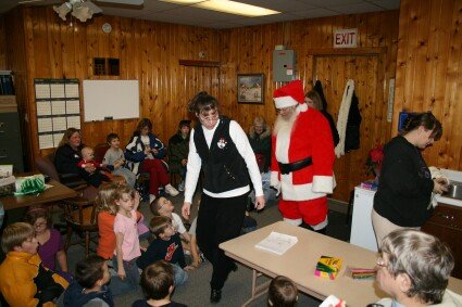 Children watch a costumed Santa and a white-clad character in a wooden community room near a table.
